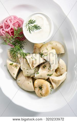 Pickles set with homemade pickled milk mushrooms, marinated red onion rings and sour cream on white restaurant plate isolated. Salted Lactarius resimus mushroom with oil and fresh chopped dill