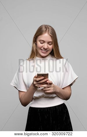 Young Pretty Blond Girl With Long Hair Smiling And Using Cell Phone Isolated On Gray Background. Con