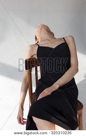 Young Beautiful Caucasian Girl In Black Dress Poses On Wooden Chair On White Emty Cyclorama In Patte
