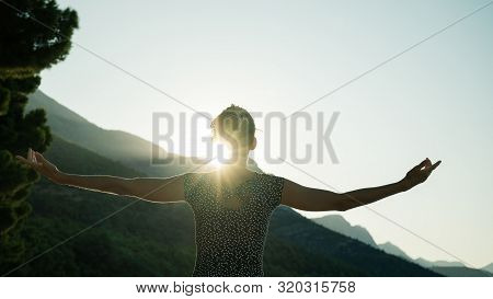 Rear View Of A Young Woman Standing To Greet Sun Rising From The Mountains  With Her Arms Wide Open.