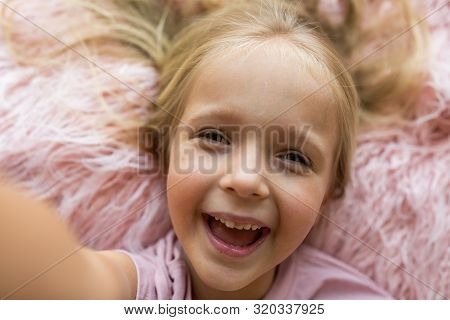 Adorable Little Girl With Long Blonde Hair Lying On Pink Fur Blanket And Making Selfie. Positive Emo