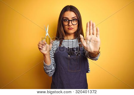 Young beautiful hairdresser woman holding scissors standing over isolated yellow background with open hand doing stop sign with serious and confident expression, defense gesture