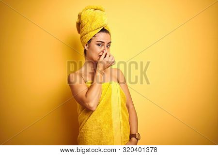 Young beautiful woman wearing towel after shower over isolated yellow background smelling something stinky and disgusting, intolerable smell, holding breath with fingers on nose. Bad smells concept.