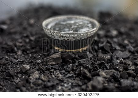 Close Up Of Activated Charcoal In A Glass Bowl On The Wooden Surface Along With Some Raw Powder Of C