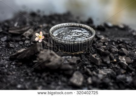 Close Up Of Activated Charcoal In A Glass Bowl On The Wooden Surface Along With Some Raw Powder Of C