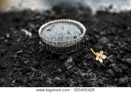 Close Up Of Activated Charcoal In A Glass Bowl On The Wooden Surface Along With Some Raw Powder Of C