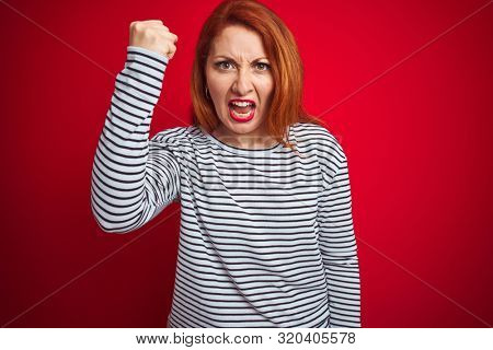Young redhead woman wearing strapes navy shirt standing over red isolated background angry and mad raising fist frustrated and furious while shouting with anger. Rage and aggressive concept.