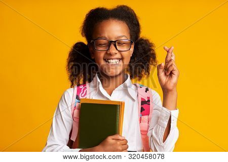 First School Day. Afro Schoolgirl Keeping Fingers Crossed For Luck Holding Books Over Yellow Studio 