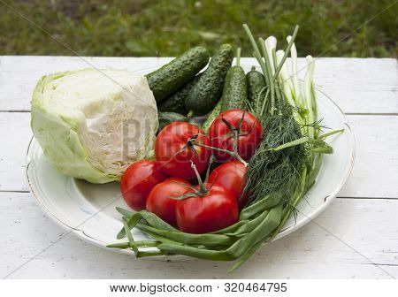 Fresh Tasty Vegetables From The Garden Lie On A Plate On A White Table. Cabbage, Cucumbers, Tomatoes