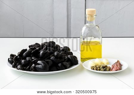 Apenade Ingredients: Pitted Black Olives On White Plate, Olive Oil In Glass Bottle, Three Cloves Of 