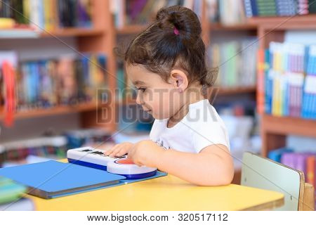 Little Child Indoors In Front Of Books. Cute Young Toddler Sitting On A Chair Near Table And Play Wi