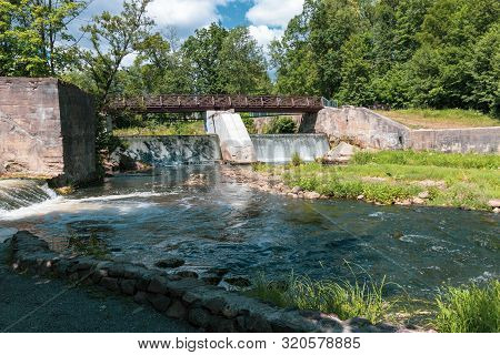 Wooden Bridge Over The River Vilia In Vilnius With A Waterfall In The Park Belmonto.