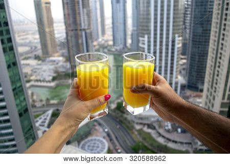 Female And Male Hands Hold Glasses Of Fresh Cold Orange Juice With City Skyscrapers View Of Modern C