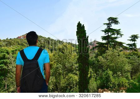 Young Travelling Man With Backpack Looks At Panoramic View Of The City In The Middle Of The Forest. 