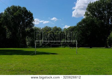 Two Football Or Soccer Goalposts In Parallel In A Park With Green Grass And Blue Skies And People Si