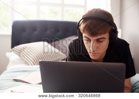 Male College Student Wearing Headphones Lies On Bed In Shared House Working On Laptop