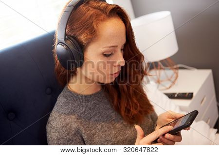 Female College Student Wearing Headphones Sitting On Bed In Shared House With Mobile Phone