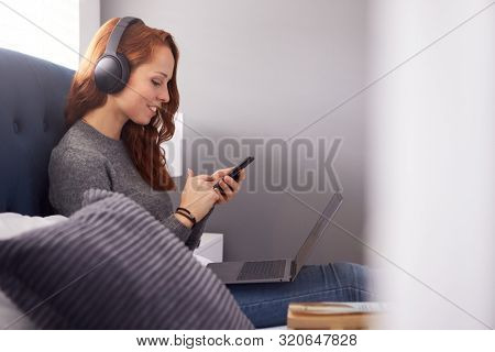 Female College Student Wearing Headphones Works On Bed In Shared House With Laptop And Mobile Phone