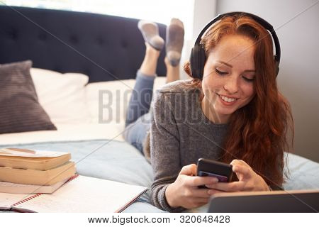 Female College Student Wearing Headphones Lies On Bed In Shared House With Mobile Phone Using Laptop