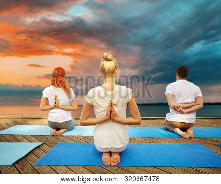fitness, sport, yoga and healthy lifestyle concept - group of people exercising in reverse prayer pose on sea pier over sunset background