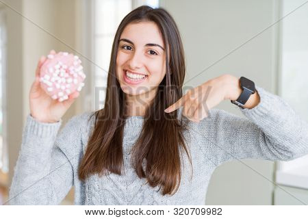 Beautiful young woman eating sugar marshmallow pink donut with surprise face pointing finger to himself