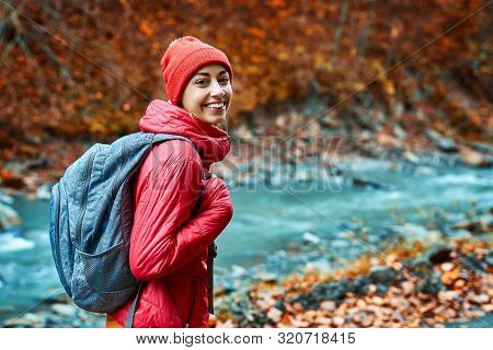 Pretty Woman Hiker In Bright Red Outdoor Clothes Is Standing Outdoors On The Mountain River And Fore