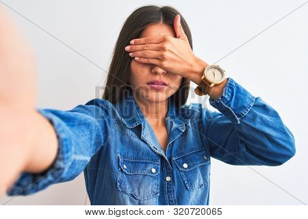 Beautiful woman wearing denim shirt make selfie by camera over isolated white background covering eyes with hand, looking serious and sad. Sightless, hiding and rejection concept