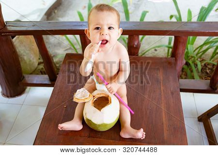 Infant Baby At Tropical Vacation. Eats And Drinks Green Young Coconut. Sits On A Wooden Table And Gn