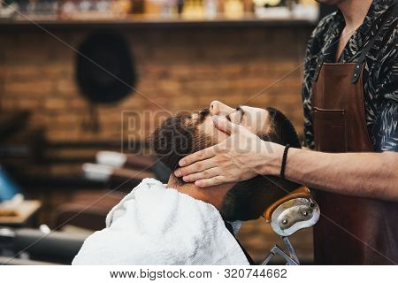 Handsome Man With A Beard And Closed Eyes In A Black Cutting Hair Cape In The Barbershop. Barber In