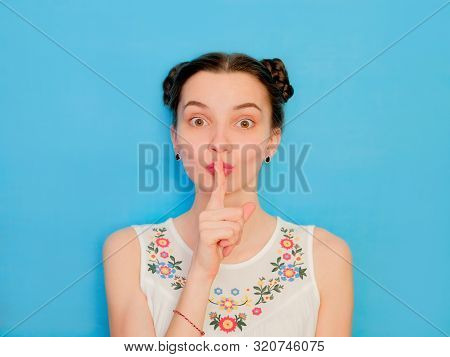 Funny Cute Girl On A Blue Studio Background. Bright Emotional Female Portrait. Secret Face Finger To