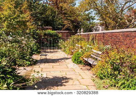 A Stone Walkway In A Garden Of Floers With Two Wooden Benches And A Brick Wall In Summer, Pittsburgh