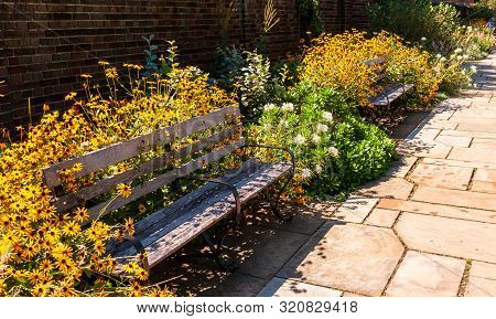 A Stone Walkway In A Garden Of Floers With Two Wooden Benches And A Brick Wall In Summer, Pittsburgh