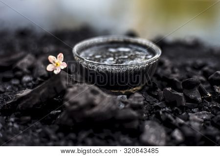 Close Up Of Activated Charcoal In A Glass Bowl On The Wooden Surface Along With Some Raw Powder Of C