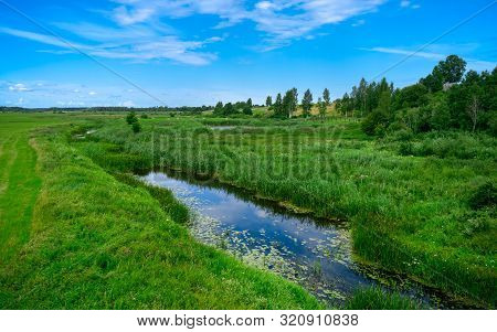 A Narrow Water Canal, River, Stream Going Through A Green Grass Field Landscape Into Bright Summer C