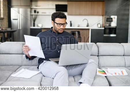 Mixed-race Indian Man In Casual Shirt Sits On The Couch And Using Laptop For Remote Work, Focused Fr