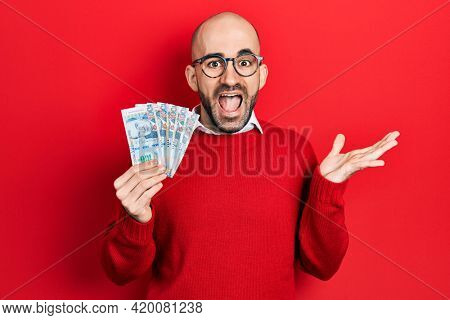 Young bald man holding peruvian sol banknotes celebrating achievement with happy smile and winner expression with raised hand 