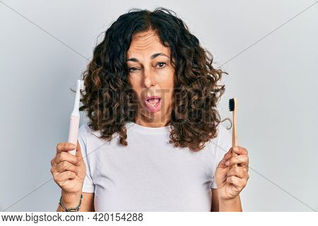 Middle age hispanic woman holding electric toothbrush and teethbrush clueless and confused expression. doubt concept. 