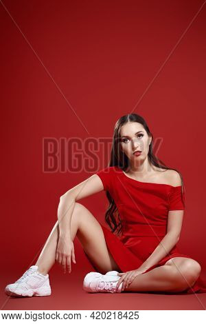 Beautiful brunette girl posing at studio in red dress and white sneakers. Red background. Sport chic style.