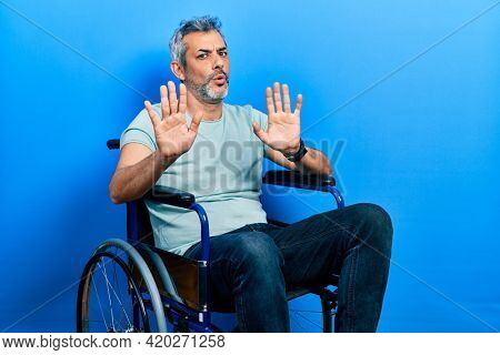 Handsome middle age man with grey hair sitting on wheelchair moving away hands palms showing refusal and denial with afraid and disgusting expression. stop and forbidden. 