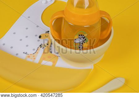 Baby Accessories On Light Yellow Background, Flat Lay. Baby Bib And Food Plate, Spoon And Baby Cup.