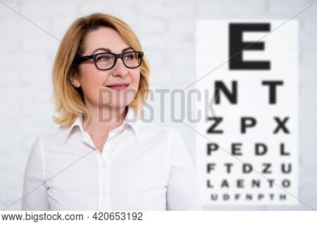 Portrait Of Thoughtful Cheerful Mature Business Woman In Eyeglasses With Eye Test Chart Over White W