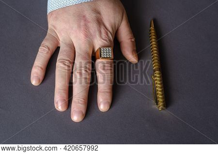A Hand With A Gold Ring And Pen On A Gray Background. The Right Hand Of An Adult Male With A Signet 