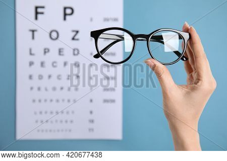 Woman Holding Glasses Against Eye Chart On Blue Background, Closeup. Ophthalmologist Prescription