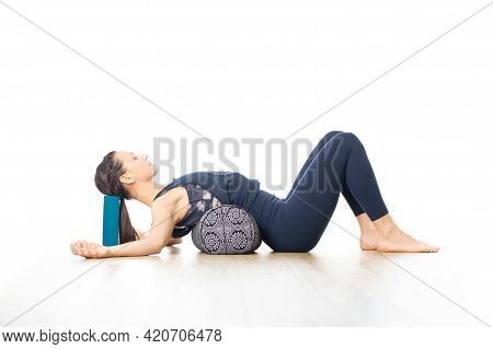 Restorative Yoga With A Bolster. Young Sporty Attractive Woman In Bright White Yoga Studio, Lying On