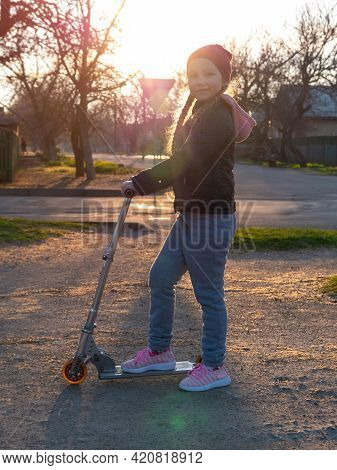 Child Riding On Kickscooter Alone. Blonde Cute Girl On Scooter On Spring City Park Street Background