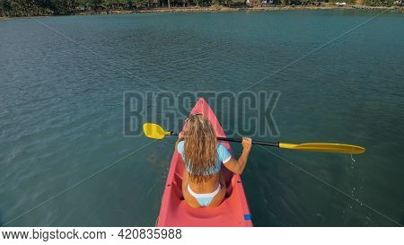 Seductive Blonde Woman In Blue Swimsuit Sails On Pink Plastic Kayak Along Azure Sea At Exotic Resort