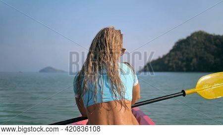Long Curly Haired Blonde Woman In Swimsuit Rows Plastic Kayak Along Azure Ocean Water At Exotic Reso
