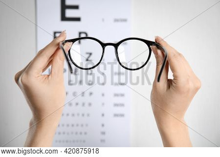 Woman Holding Glasses Against Eye Chart On Light Background, Closeup. Ophthalmologist Prescription