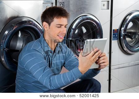 Young man using digital tablet while sitting against washing machines at laundry