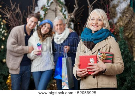 Portrait of happy senior woman holding Christmas presents with family standing in background at store